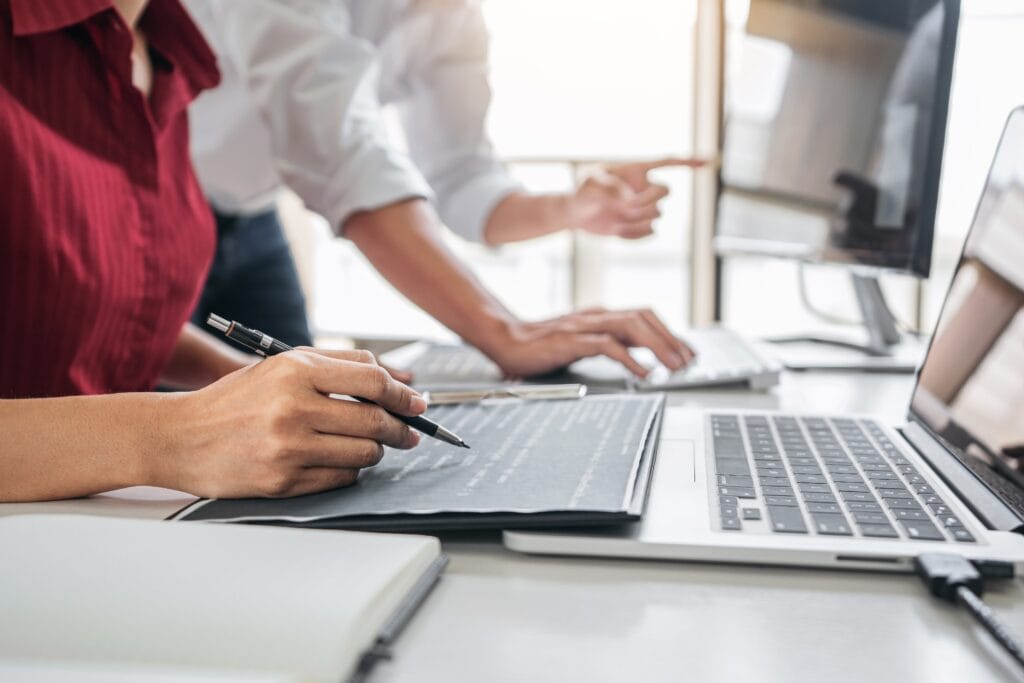 The image depicts a professional setting where two individuals appear to be collaborating on a project. On the right, one person is pointing towards a computer screen, perhaps giving instructions or explaining a concept. This individual seems to be wearing a white shirt, which suggests a business casual attire commonly seen in office environments. On the left, another person is seated and appears to be taking notes or writing something down, indicating active participation in the discussion or task at hand. They are wearing a red top, which adds a pop of color to the image. In front of them are multiple laptops, signifying a tech-centric workspace. The laptops are open, indicating that work is in progress. The presence of multiple laptops suggests that the task at hand may involve coding, data analysis, or other computer-related activities, often associated with programming or machine learning projects. The notebooks and pens on the table further suggest that this is a brainstorming session or a collaborative meeting where ideas are being discussed and recorded. The overall atmosphere conveyed by the image is one of focused teamwork, with an emphasis on technology and collaboration, typical of a machine learning or programming environment.
